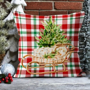 May include: A red and green plaid throw pillow with a Christmas tree and presents in a wooden sled. The tree is decorated with lights and the presents are wrapped in white paper with brown ribbon.