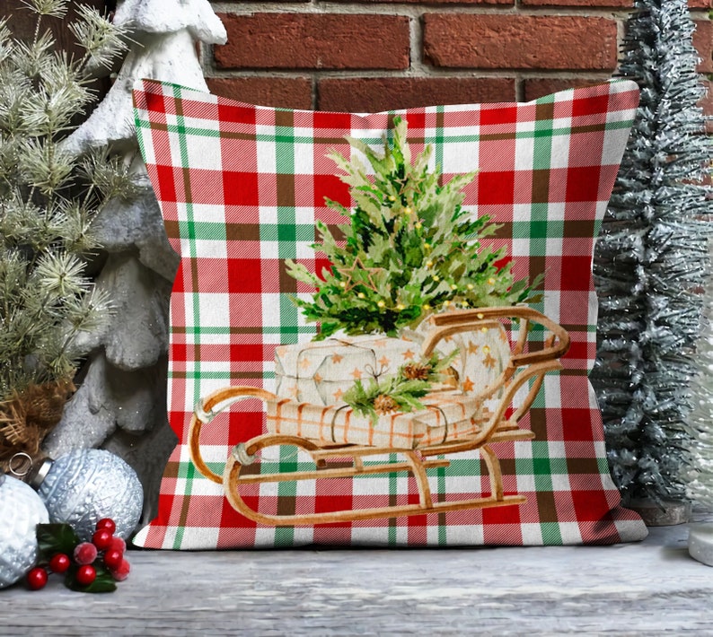 May include: A red and green plaid throw pillow with a Christmas tree and presents in a wooden sled. The tree is decorated with lights and the presents are wrapped in white paper with brown ribbon.