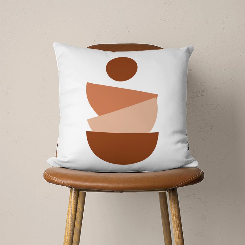 Pu&ograve; includere: Un cuscino bianco con un design geometrico che presenta tre forme semicircolari impilate in tonalit&agrave; di marrone, arancione e pesca. Le forme sono centrate sul cuscino.
