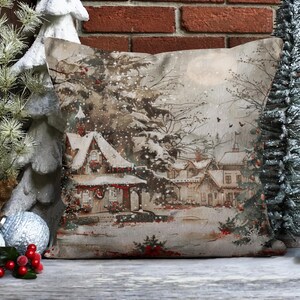 May include: A decorative pillow with a winter scene featuring a snow-covered village with houses and trees. The pillow has a beige background with a brown and white pattern.