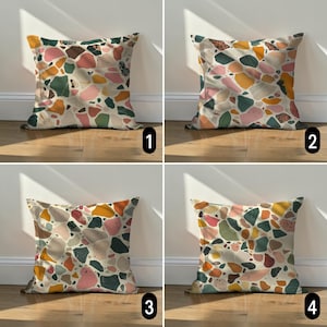 Pode incluir: Quatro almofadas quadradas com um padrão de terrazzo. Cada almofada apresenta uma disposição diferente de formas coloridas em tons de rosa, verde, laranja e castanho sobre um fundo creme. As almofadas são exibidas num chão de madeira.