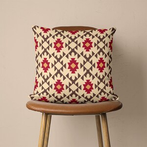 Op de afbeelding: Een decoratief kussen met een geometrisch patroon in beige, bruin, rood en oranje tinten. Het kussen heeft een herhalend ontwerp van driehoeken en vierkanten.