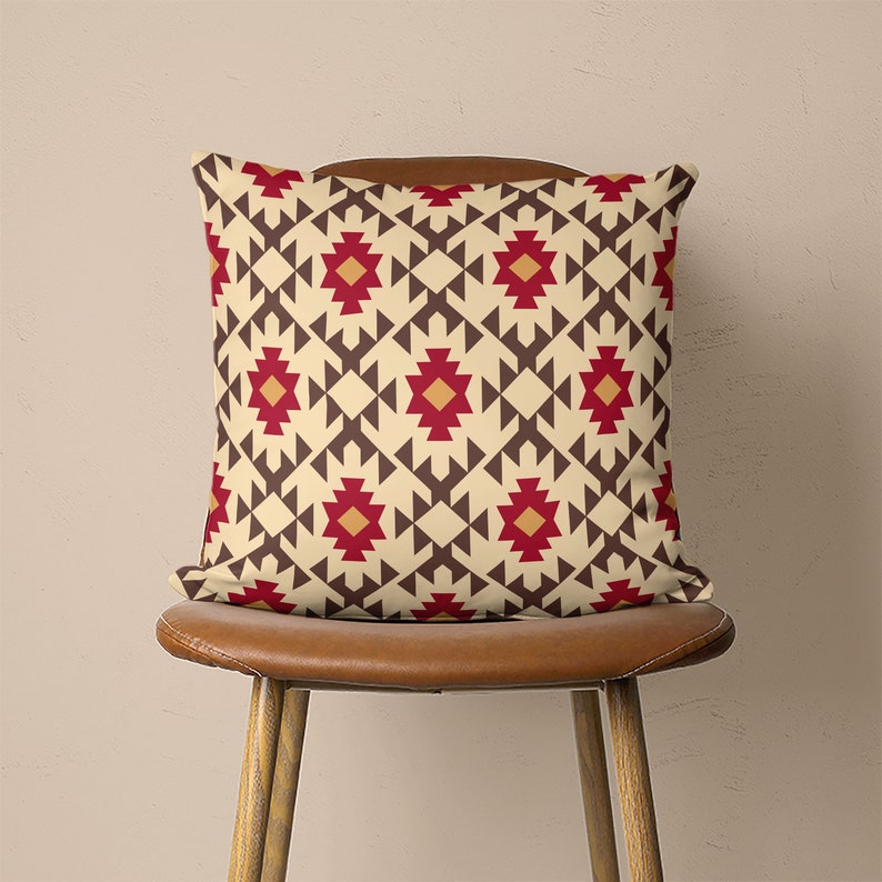 Op de afbeelding: Een decoratief kussen met een geometrisch patroon in beige, bruin, rood en oranje tinten. Het kussen heeft een herhalend ontwerp van driehoeken en vierkanten.