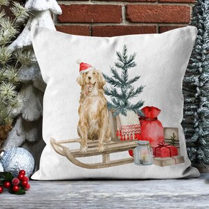 May include: A white throw pillow with a watercolor illustration of a golden retriever wearing a Santa hat, riding a wooden sled with a small Christmas tree, a red bag, and a glass jar with a candle.