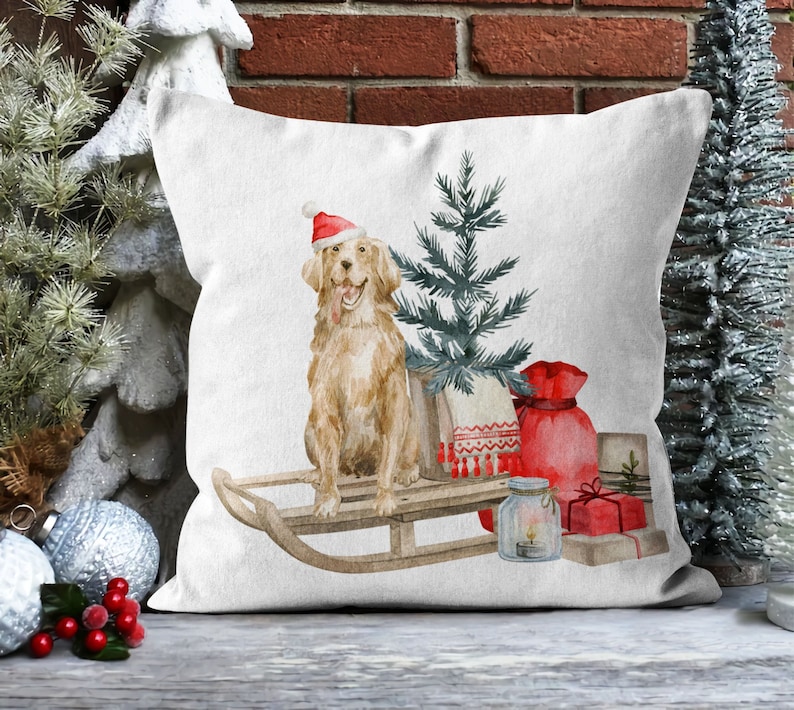 May include: A white throw pillow with a watercolor illustration of a golden retriever wearing a Santa hat, riding a wooden sled with a small Christmas tree, a red bag, and a glass jar with a candle.