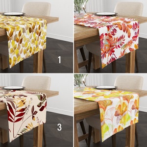 May include: Four table runners featuring different autumn leaf designs. Each runner is white with a variety of yellow, orange, and red leaves. The runners are displayed on wooden tables with a white chair in the background.