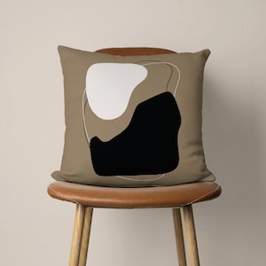Puede incluir: Una almohada marr&oacute;n con un dise&ntilde;o abstracto en blanco y negro. La almohada est&aacute; sentada en una silla de cuero marr&oacute;n.