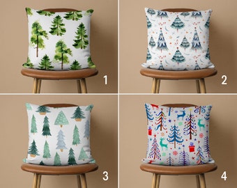 May include: Four different Christmas pillow covers with various designs. The first pillow cover features a watercolor design of green Christmas trees on a white background. The second pillow cover features a blue and white Christmas tree design with red berries and stars. The third pillow cover features a blue and green Christmas tree design with a white background. The fourth pillow cover features a colorful Christmas tree design with red, blue, green, and white colors. The pillow covers are all on brown chairs.
