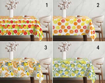 May include: Four different tablecloths with citrus fruit designs. The tablecloths are white with various patterns of lemons, limes, and oranges. The tablecloths are shown on a table with a white chair.