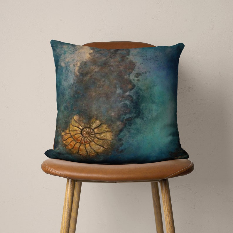 May include: A teal blue throw pillow with a watercolor design featuring a fossilized seashell in shades of gold and brown. The pillow is sitting on a brown leather chair.
