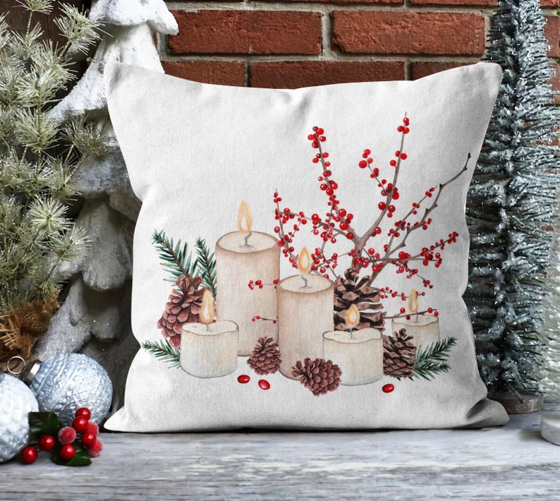 May include: A white throw pillow with a wintery design featuring candles, pine cones, and red berries. The candles are white and have a warm glow. The pine cones are brown and the berries are red.