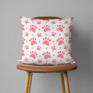 May include: Square white pillow with a repeating pattern of pink paw prints and small hearts. The pillow is placed on a brown leather chair. The design is a watercolor style.