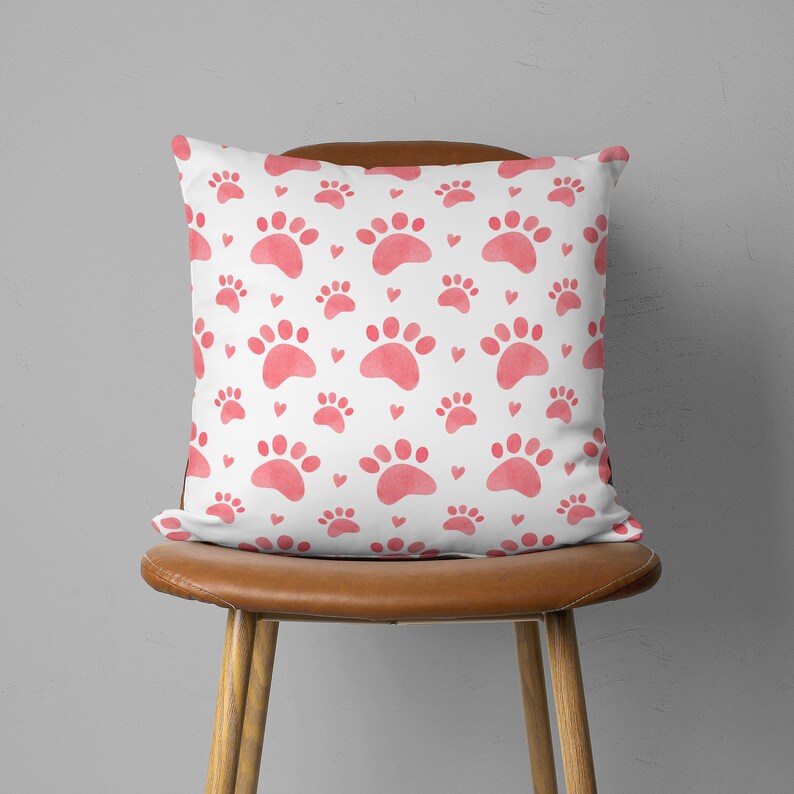 May include: Square white pillow with a repeating pattern of pink paw prints and small hearts. The pillow is placed on a brown leather chair. The design is a watercolor style.