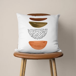 Puede incluir: Almohada blanca con un dise&ntilde;o abstracto marr&oacute;n, dorado y naranja con un patr&oacute;n de puntos negros.