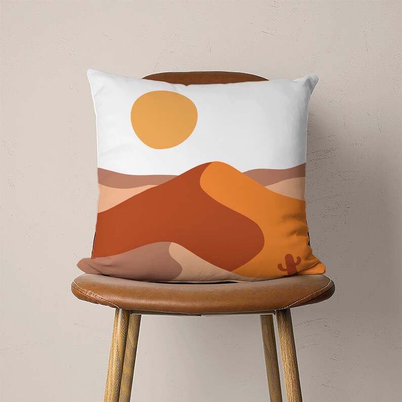 Pu&ograve; includere: Un cuscino bianco con un design a paesaggio desertico. Il cuscino presenta una grande duna arancione e marrone con un piccolo sole giallo nel cielo. Un piccolo cactus si trova in primo piano.