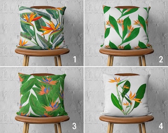Op de afbeelding: Vier kussens met verschillende ontwerpen met tropische paradijsvogelbloemen op een witte achtergrond. De kussens worden getoond op bruine houten stoelen tegen een witte bakstenen muur.