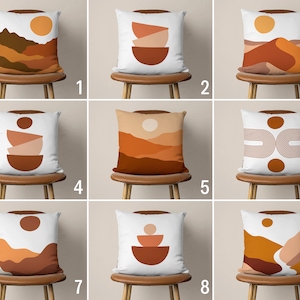 Pu&ograve; includere: Nove diversi design di cuscini con uno sfondo bianco e vari motivi astratti in tonalit&agrave; di marrone, arancione e giallo. I cuscini sono esposti su sedie in legno con una finitura marrone chiaro.