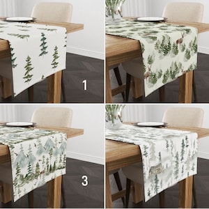 May include: Four different table runners featuring woodland-themed designs. The runners are all white with green and brown watercolour illustrations of trees, deer, and a cabin. The runners are displayed on wooden tables against a white backdrop.