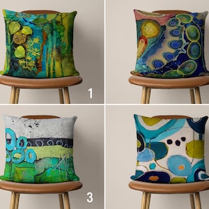 Pode incluir: Quatro almofadas decorativas com designs abstratos em tons de azul, verde e amarelo. As almofadas são exibidas em cadeiras marrons com pernas de madeira.