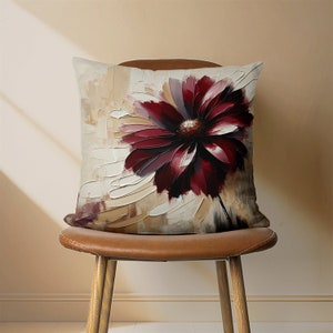 May include: A square throw pillow with a cream background and a large, abstract burgundy flower design. The pillow is sitting on a brown leather chair.