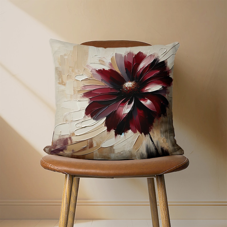 May include: A square throw pillow with a cream background and a large, abstract burgundy flower design. The pillow is sitting on a brown leather chair.