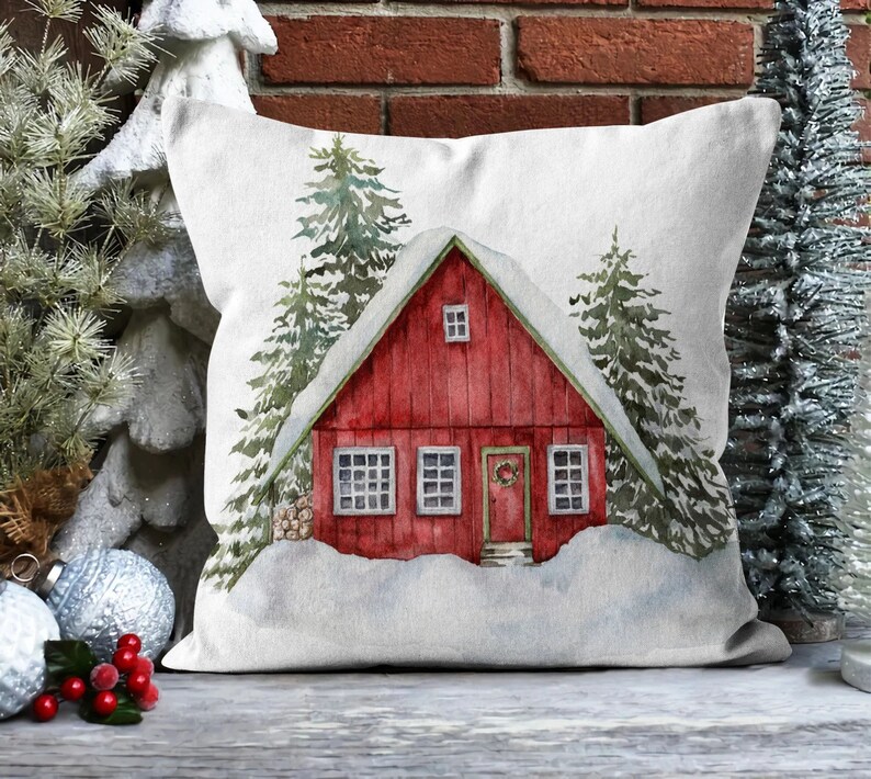 Op de afbeelding: Een wit kussen met een rood huisje in een besneeuwde winterse sc&egrave;ne. Het huisje heeft een krans op de deur en er staan besneeuwde bomen omheen.