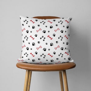 May include: White square throw pillow with a pattern of black paw prints and red and white striped dog bones. The pillow is on a brown leather chair.