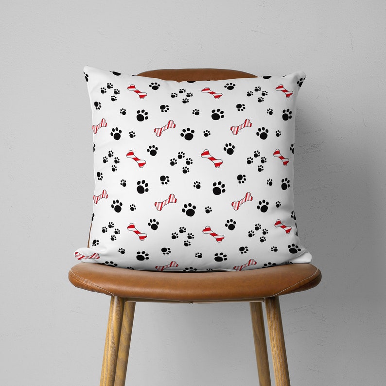 May include: White square throw pillow with a pattern of black paw prints and red and white striped dog bones. The pillow is on a brown leather chair.