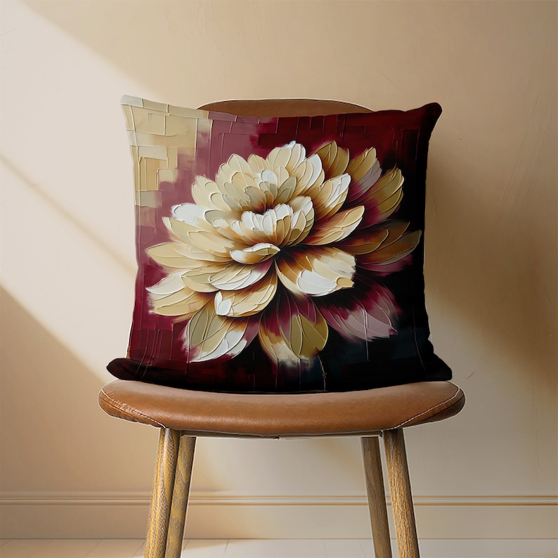 May include: A decorative throw pillow with a large, white and gold flower design on a burgundy background. The pillow is sitting on a brown leather chair.