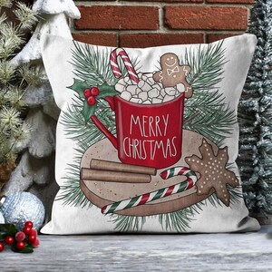 Op de afbeelding: Een wit kussen met een rode mok gevuld met marshmallows en een peperkoekman. De mok zegt "Merry Christmas". Er zijn ook kaneelstokjes en een snoepstok op een bruin bord.
