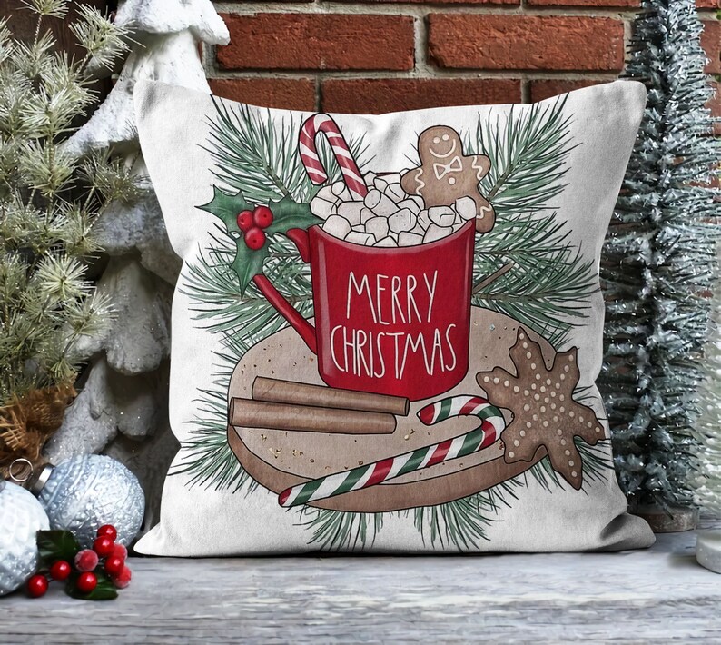 Op de afbeelding: Een wit kussen met een rode mok gevuld met marshmallows en een peperkoekman. De mok zegt "Merry Christmas". Er zijn ook kaneelstokjes en een snoepstok op een bruin bord.