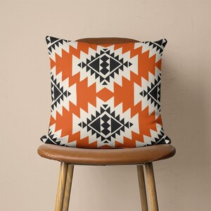 Op de afbeelding: Een oranje en zwart geometrisch gepatroneerd kussen met een witte achtergrond. Het kussen ligt op een bruine stoel.