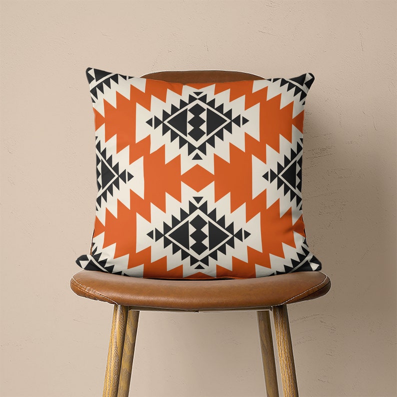 Op de afbeelding: Een oranje en zwart geometrisch gepatroneerd kussen met een witte achtergrond. Het kussen ligt op een bruine stoel.