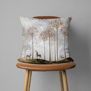 May include: A decorative throw pillow with a winter forest scene featuring brown and white deer silhouettes and trees with orange leaves against a gray and white background.