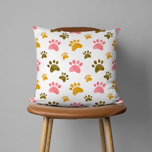 May include: Square white pillow with a repeating pattern of paw prints in pink, gold, and brown. The pillow is on a brown leather chair with wooden legs. The paw prints vary in size.