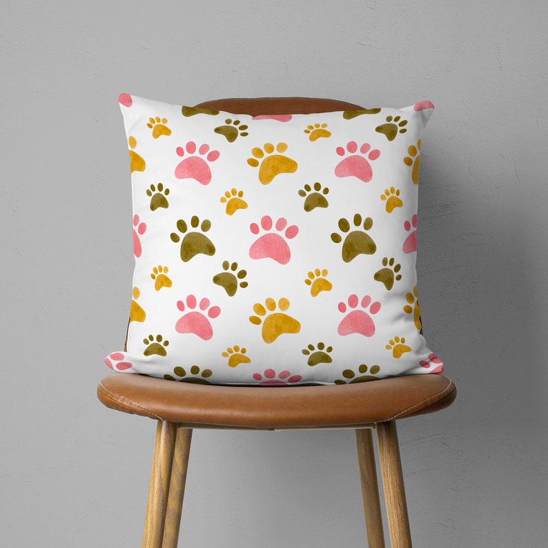 May include: Square white pillow with a repeating pattern of paw prints in pink, gold, and brown. The pillow is on a brown leather chair with wooden legs. The paw prints vary in size.