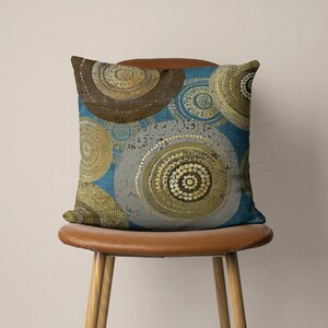 Puede incluir: Una almohada decorativa con un patr&oacute;n geom&eacute;trico azul, gris y dorado. La almohada presenta un dise&ntilde;o circular con c&iacute;rculos conc&eacute;ntricos y detalles intrincados.