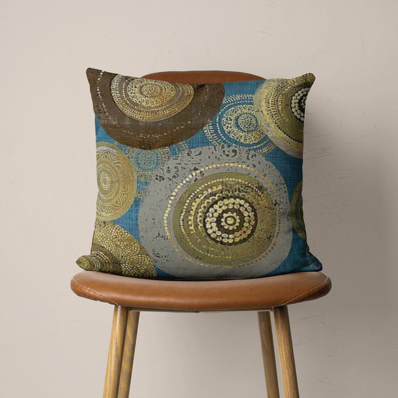 Puede incluir: Una almohada decorativa con un patr&oacute;n geom&eacute;trico azul, gris y dorado. La almohada presenta un dise&ntilde;o circular con c&iacute;rculos conc&eacute;ntricos y detalles intrincados.