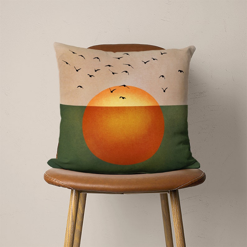 May include: A green and beige throw pillow with a graphic of a large orange sun setting over a green field. Silhouettes of birds fly across the sky above the sun.