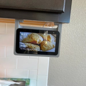May include: A gray Amazon Echo Show smart display mounted under a kitchen cabinet. The screen displays a recipe for biscuits with the text "Popular Recipes" and "Biscuits". The time is 8:21 and the temperature is 30 degrees.