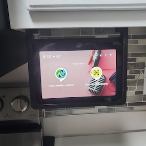 May include: A black tablet mounted under a kitchen cabinet with a pink screen displaying the Google Assistant interface. The screen shows the time 5:53 and the temperature 36 degrees. The text "Open ambient visions" and "Open fan sounds" are displayed on the screen.