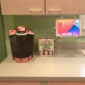 May include: A pink and black Keurig coffee maker with a strawberry design sits on a white countertop next to a white ceramic holder with four cups. The holder has a graphic of a diner with the text "Sophia's Diner". A white tablet is mounted on the wall above the countertop.
