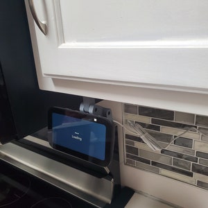 May include: A black and gray smart home device with a blue screen that says "Loading..." is mounted under a white cabinet. The device is connected to an outlet with a white cord.