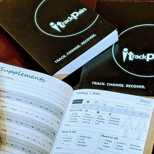 May include: Two black notebooks with the text "iTrackPain" and "TRACK. CHANGE. RECOVER." on the covers. One notebook is open to a page with a section for tracking supplements and another section for tracking pain, sleep, and mood.