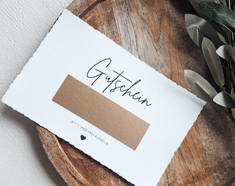 May include: A white card with the word "Gutschein" written in black cursive. The card has a brown scratch-off area with the text "Bitte hier frei rubbeln" and a small black heart.