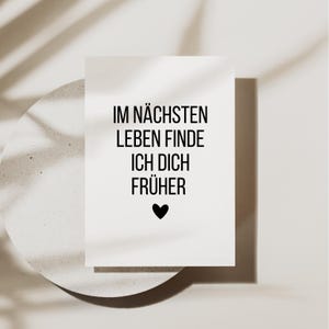 May include: A white card with black text that reads "IM N&Auml;CHSTEN LEBEN FINDE ICH DICH FR&Uuml;HER" and a black heart. The card is on a light-colored, textured surface with a shadow of a leaf.