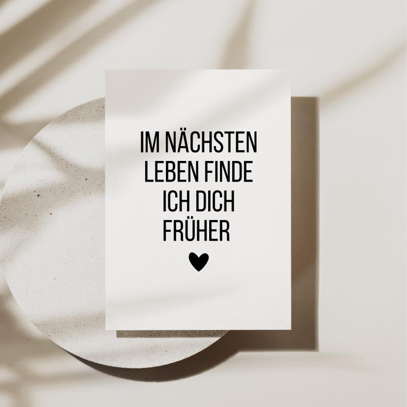May include: A white card with black text that reads "IM N&Auml;CHSTEN LEBEN FINDE ICH DICH FR&Uuml;HER" and a black heart. The card is on a light-colored, textured surface with a shadow of a leaf.