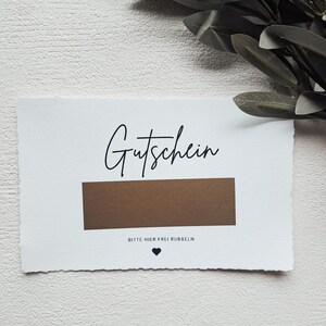 May include: A white card with the word "Gutschein" written in black cursive. The card has a brown scratch-off area with the text "Bitte hier frei rubbeln" (Please rub here freely) in black text below it.