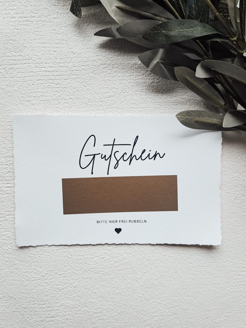 May include: A white card with the word "Gutschein" written in black cursive. The card has a brown scratch-off area with the text "Bitte hier frei rubbeln" (Please rub here freely) in black text below it.