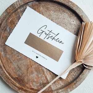 May include: A white card with the word "Gutschein" written in black cursive. The card has a gold rectangle with the text "Bitte hier frei rubbeln" written in black below it.